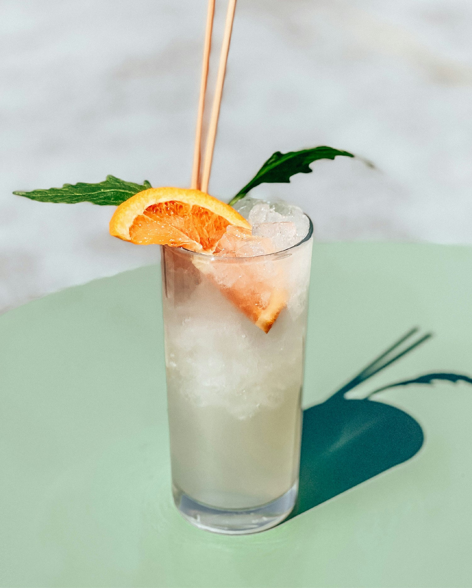 A glass of Sparkling Elixir, over ice with a fresh herb and orange wedge garnish, sitting on a light green table.