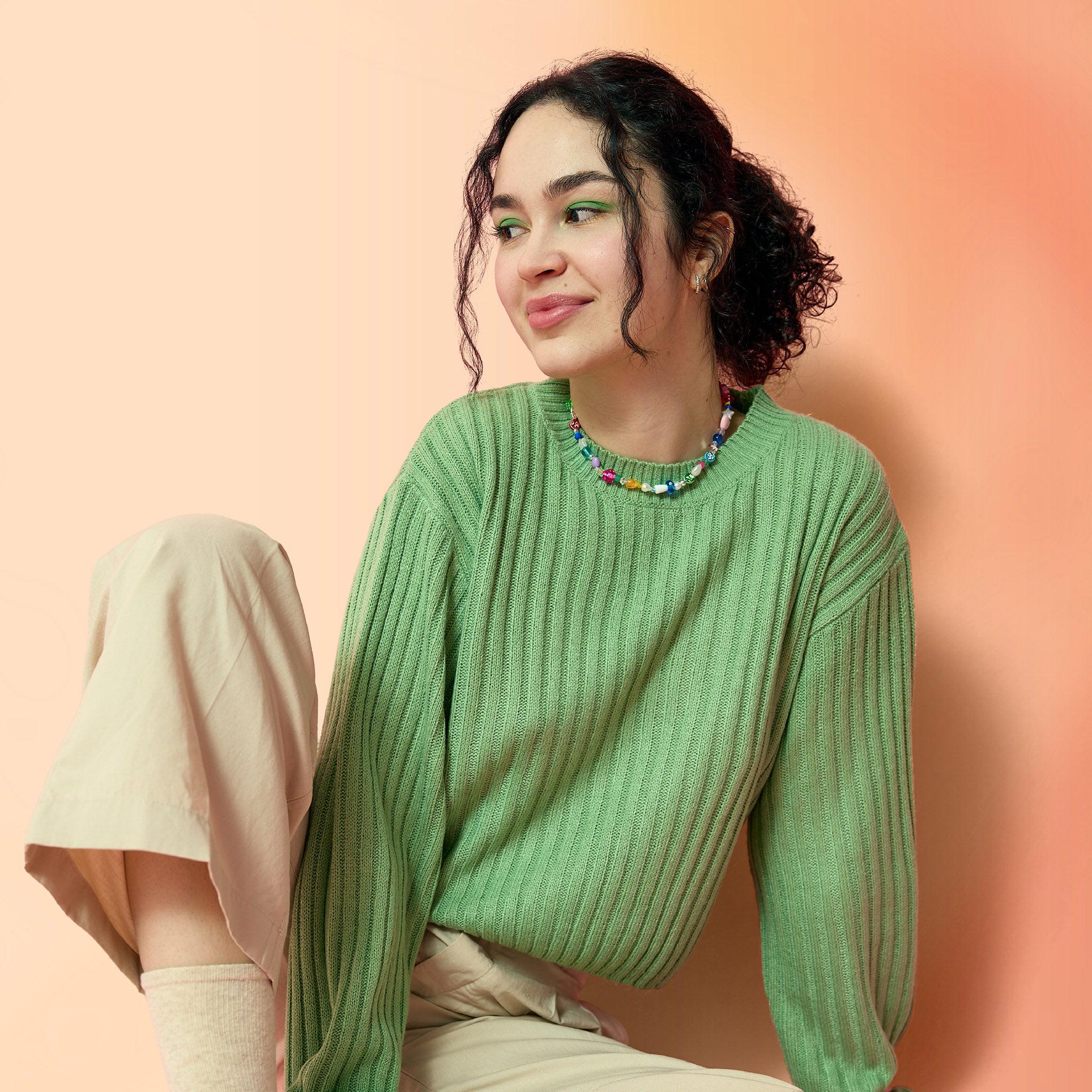 Young woman with a curly ponytail and green eyeshadow, against a pink-orange background.