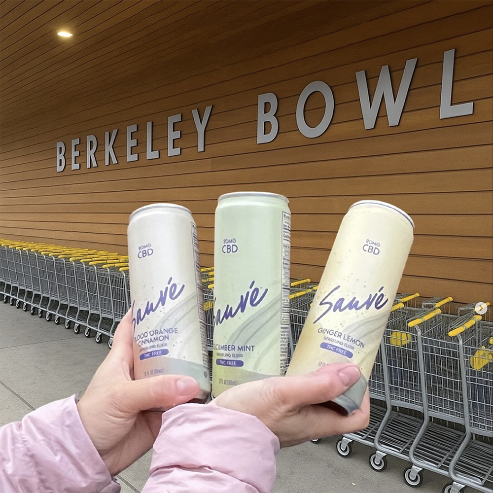 Cans of all three flavors of Sauve Sparkling Elixirs held up in front of a Berkeley Bowl sign.